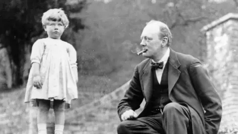 Churchill Archives Centre, Churchill Press Photographs, CHPH 1A/F2/8 A black and white photograph of Winston Church in 1924, wearing a three piece suit and bow tie, smoking a cigar, sitting on garden steps, looking at his young daughter Mary, Chartwell, Kent