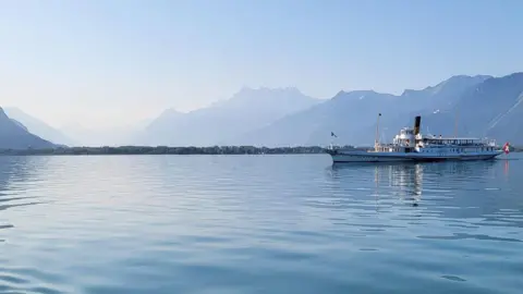 Sam Farrow/PA Lake Geneva surrounded by mountains. A boat on the right of the pic has a Switzerland flag. 