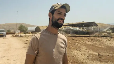 Matthew Cassel / BBC Moshe Sharvit, who has a beard and is wearing a khaki t-shirt and cap, captured in conversation with our interviewer. Behind him are metal constructions on his outpost and dusty-looking land, with a car parked behind a metal fence.