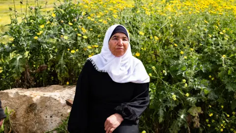 Matthew Cassel / BBC Palestinian grandmother Ayesha Shtayyeh, wearing a long-sleeved black tunic and white headscarf, with a serious expression on her face. She  sits in front of a huge swathe of yellow flowers. 