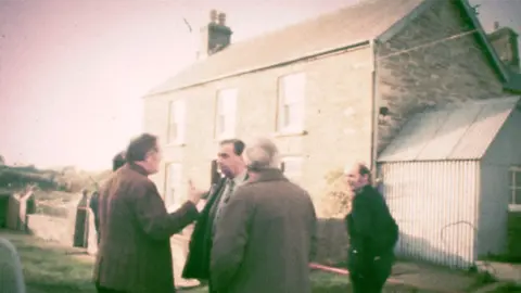 BBC News A still from archive footage of police officers outside Ffynon Samson farmhouse in December 1976. DCS Pat Malloy is stood in the centre of the group, wearing a suit and talking to his colleagues, as a uniformed officer looks on.