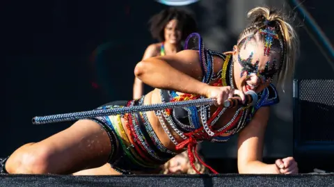 Getty Images JoJo Siwa lying on a stage singing and performing at music festival Mighty Hoopla in London in 2024. She's wearing a black and brightly-coloured outfit covered in silver chains and holding a matching microphone. She has dark eye make-up which is surrounded by multi-coloured jewels.