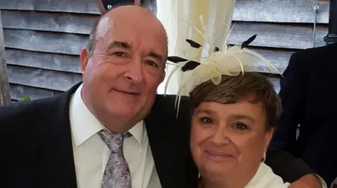 Contributed Rodrick Lodge wearing a suit has his arm around his wife Pauline who is wearing a black and white fascinator. They are both smiling and looking directly at the camera. 