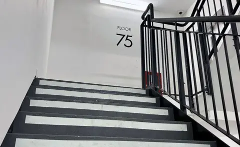 A view of the staircase at the Landmark Pinnacle building with a sign on the wall at the top of the stairs saying 'floor 75', the stairs are black and white with metallic bannisters and the walls are white.