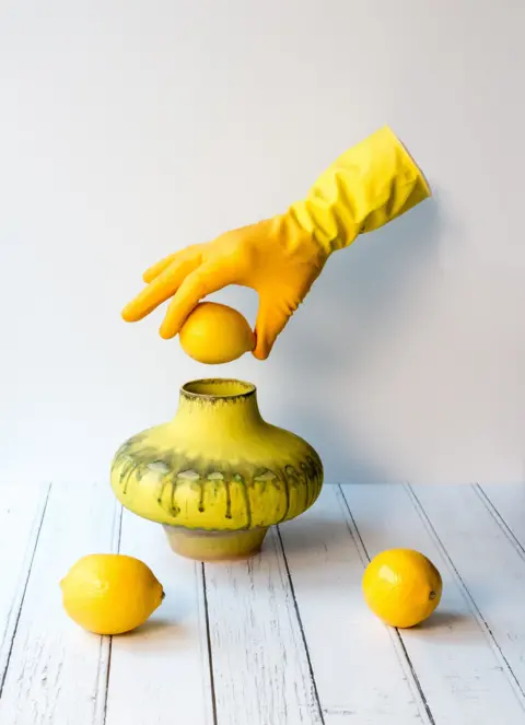 Tom Plucinski A yellow gloved hand, suspended in the air without being attached to a body, holds a lemon above a yellow vase. Two more lemons sit on a table in front of the vase.