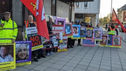 Stuart Woodward/BBC A group of at least 11 campaigners from "Matthew's campaign" standing outside an inquest in Essex. They are holding up flags and placards that show the names and photos of family members who have died. At the top of many placards reads "failed by the state".