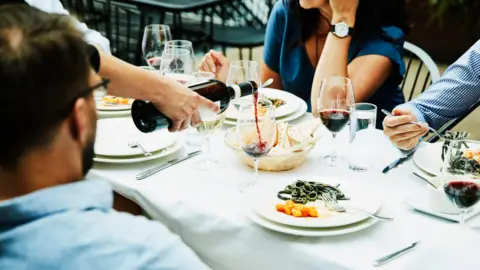 Getty Images Dining table with red wine being served. There is a white linen table cloth and finished plates of food on white plates.