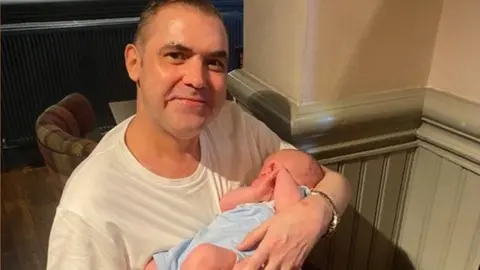 Family photograph A man with short dark hair wearing a white T-shirt smiles while he holds a baby in a blue outfit.