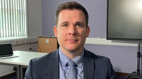 Ben Schofield/BBC Graham Newton, pictured looking direct to camera in an office, with a purple wall, part of a large TV screen and a window covered by a set of vertical blinds behind him. Mr Newton is wearing a blue shirt, dark blue and white patterned shirt and dark blue blazer. He has blue eyes, brown hair brushed to the side and a hint of stubble on his face. 