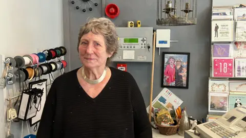 BBC Maureen with short grey hair wearing a black jumper standing behind the till of her gift shop 