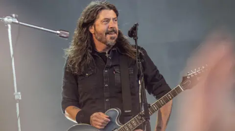 Getty Images US musician Dave Grohl of rock band Foo Fighters performs on the Orange Stage at Roskilde Festival on 5 July in Roskilde, Denmark. Grohl is wearing a black shirt and has his right hand on the top of his guitar as he poses at the front of the gig. Grohl is looking out to the crows.