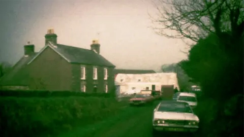 BBC News Archive image from 1976 of a grey farmhouse on a lane, with a white barn next to it and lots of old police cars parked outside