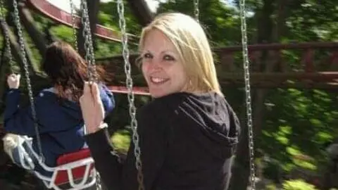 Tom Sisley Gemma Sisley is sitting on a swing, her head is turned towards the camera and she is smiling. She is wearing a black hoody.