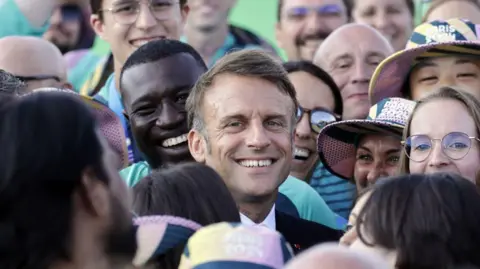 ANDRE PAIN/POOL/AFP  French President Emmanuel Macron poses with volunteers as he visits archery competitions during the Paris 2024 Olympic Games at the Esplanade des Invalides in Paris on August 2, 2024