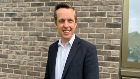 BBC Tom Pursglove with short fair hair wearing a dark jacket and  white shirt in front of a brick wall.