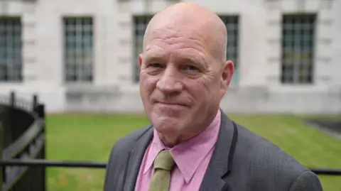 Ann Gannon / BBC Stephen Close smiles at the camera infront of a green grassy lawn, black railings and a light-coloured-brick building in Whitehall, central London. He wears a pink shirt, sage green tie and a grey suit.