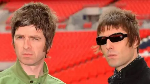 PA Media Noel Gallagher in a light green jacket and his brother, Liam, in shades, to his right with a lack jacket and black and white cravat 