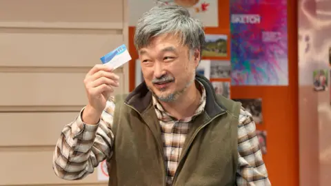 Mark Douet Ins Choi performing in Kim's Convenience, holding up a business card