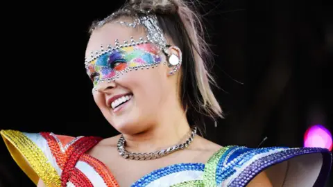 Getty Images JoJo Siwa smiling while performing on stage wearing a rainbow-coloured outfit with large shoulder pads. She's wearing multi-coloured eye make-up which is framed by silver jewels and has a matching necklace around her neck.